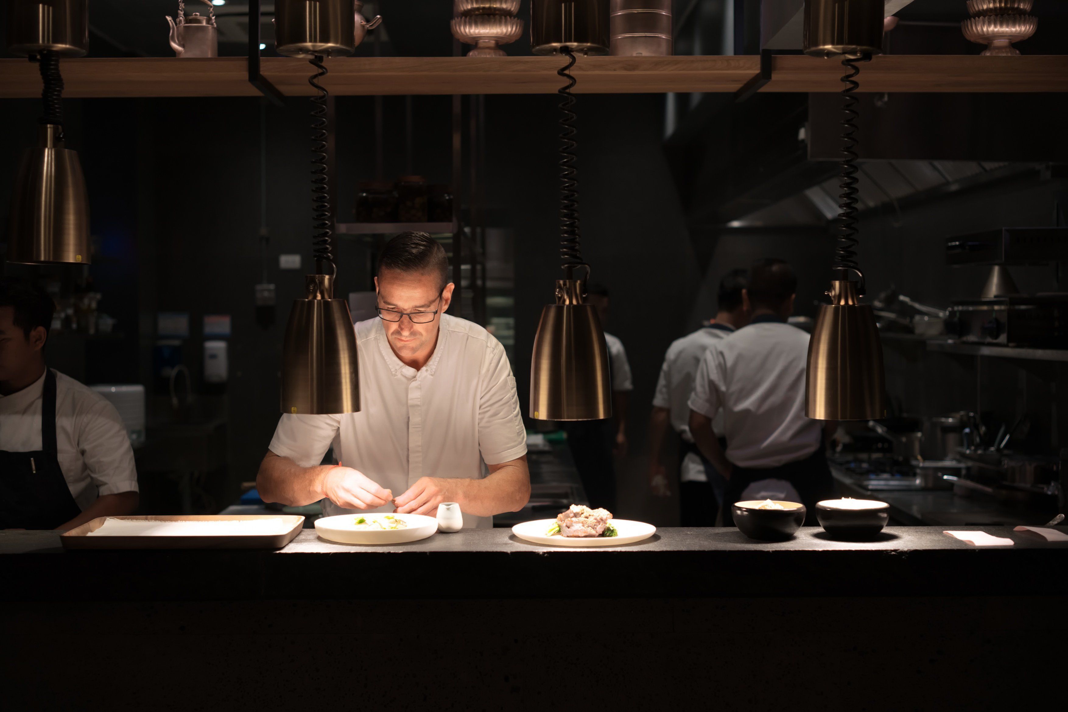 A chef preparing food in a dimly lit kitchen with hanging pendant lights.