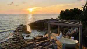 Une terrasse en bois avec une grande baignoire et une pergola en bois offrant une vue sur l’océan au coucher du soleil.