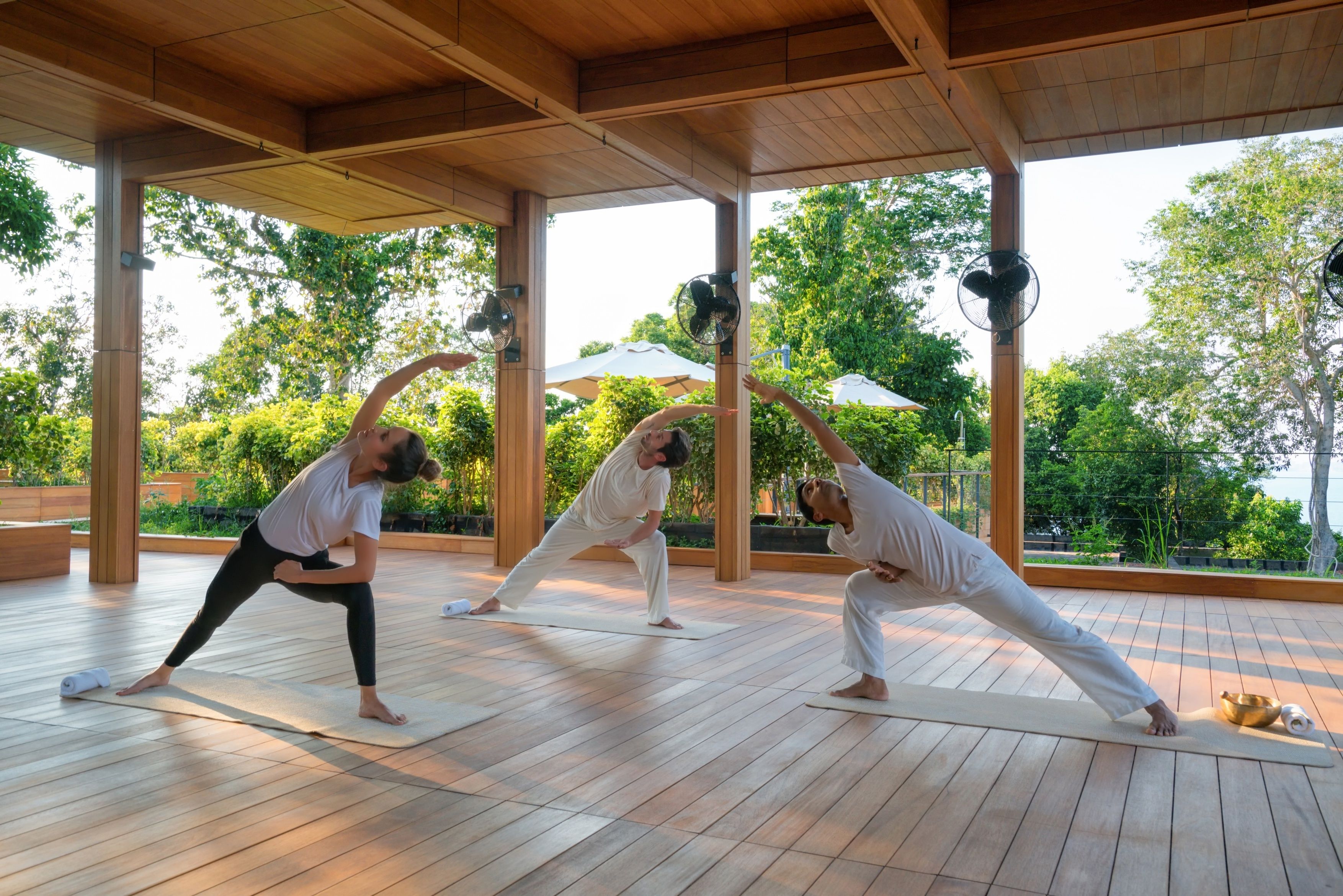 A yoga class in a wooden-floored room with large windows and fans.