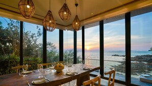 A dining area with a wooden table set for four, large windows with a view of the sea, and wicker pendant lights. - Six Senses Krabey Island (Prey Nob)