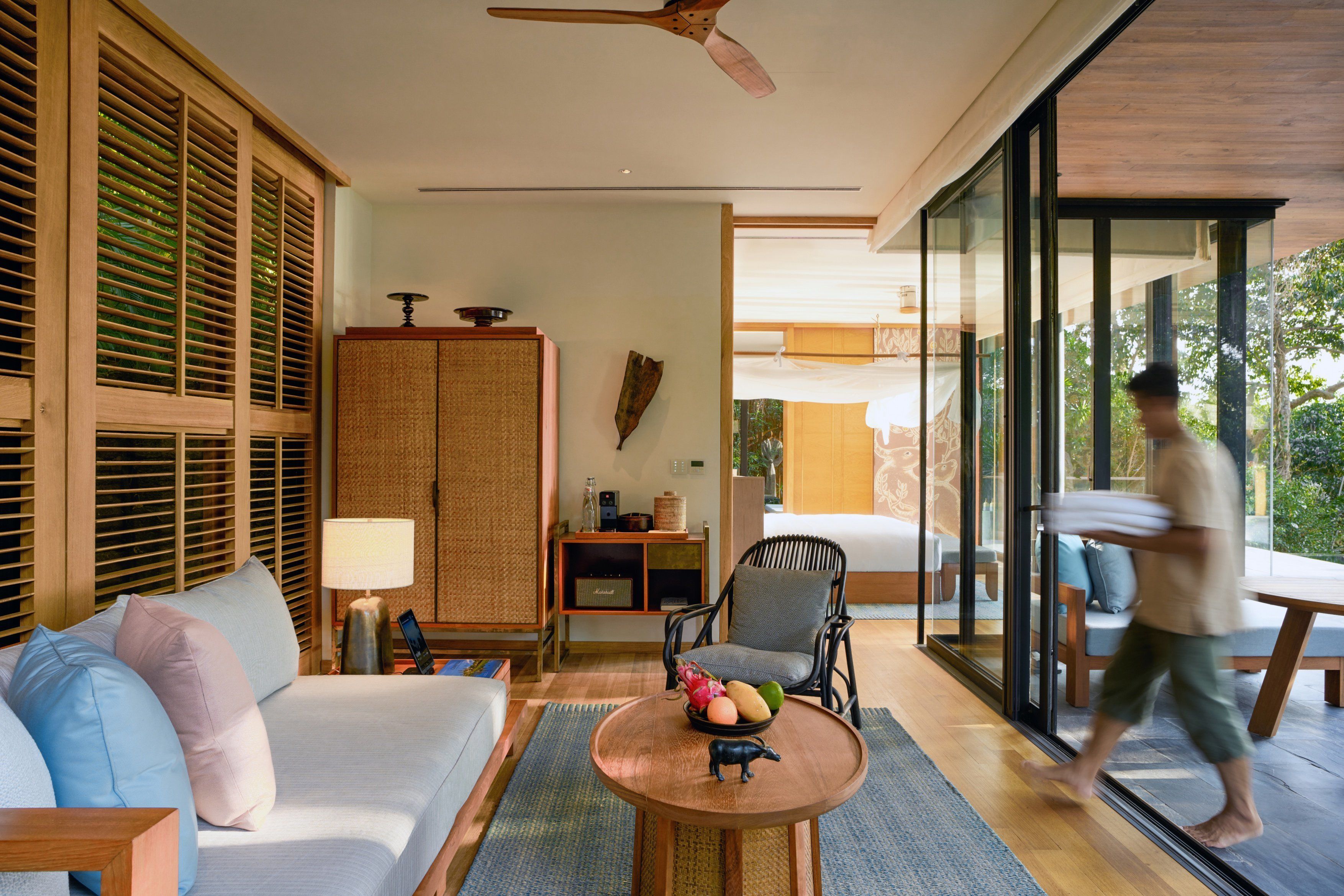 A modern hotel room with a sofa, a round wooden table with fruit, a chair, and a large wardrobe.