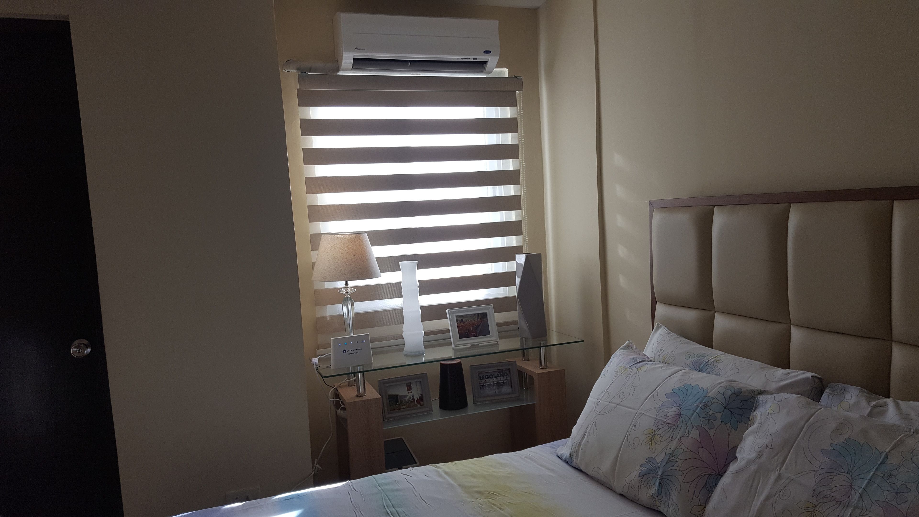 Une chambre à coucher avec un lit, une table de chevet, une lampe, un cadre photo et une fenêtre avec des stores.