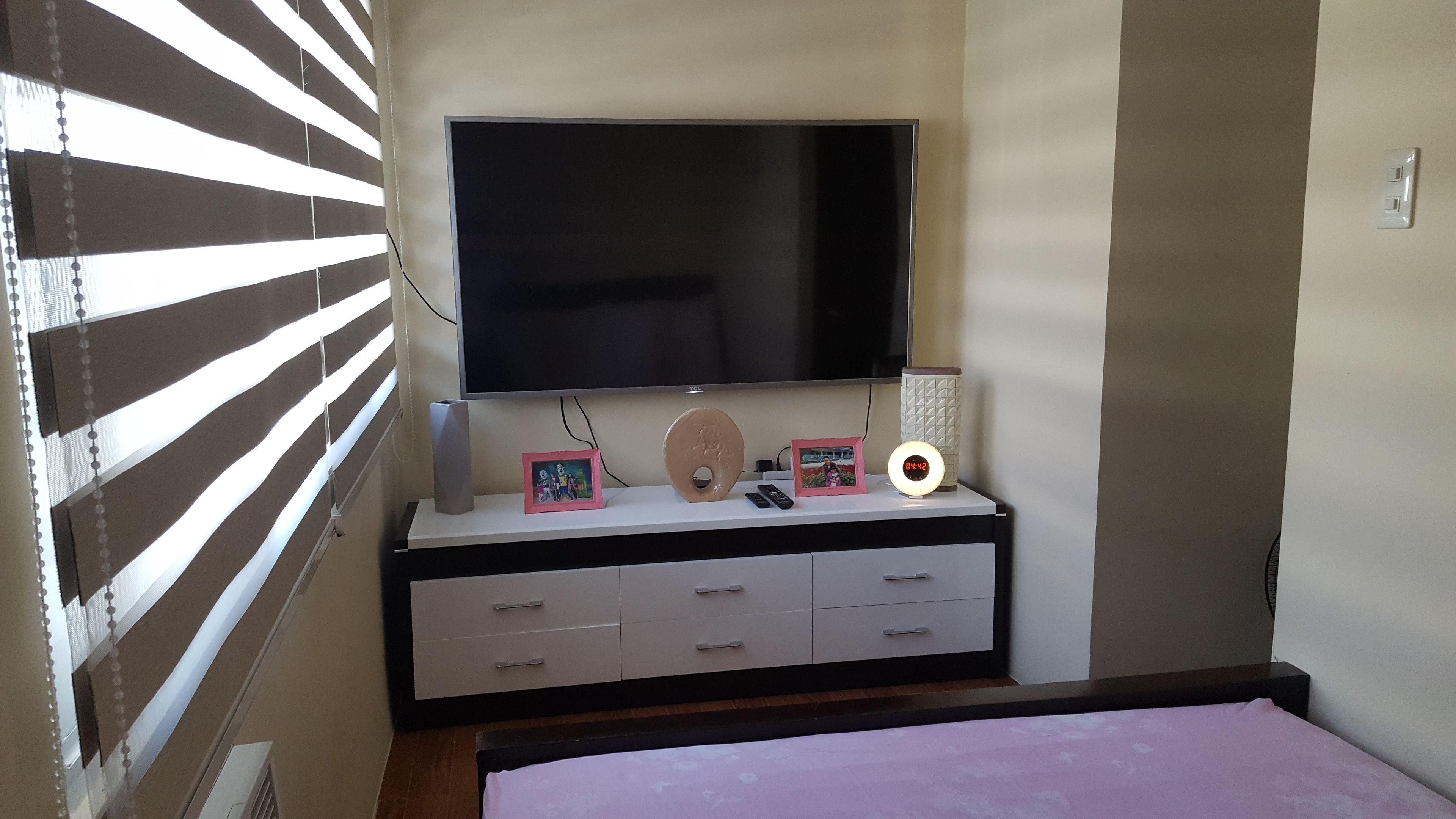 A bedroom with a TV on a white dresser, a bedside table with a lamp and a clock, and a bed with a pink bedspread.