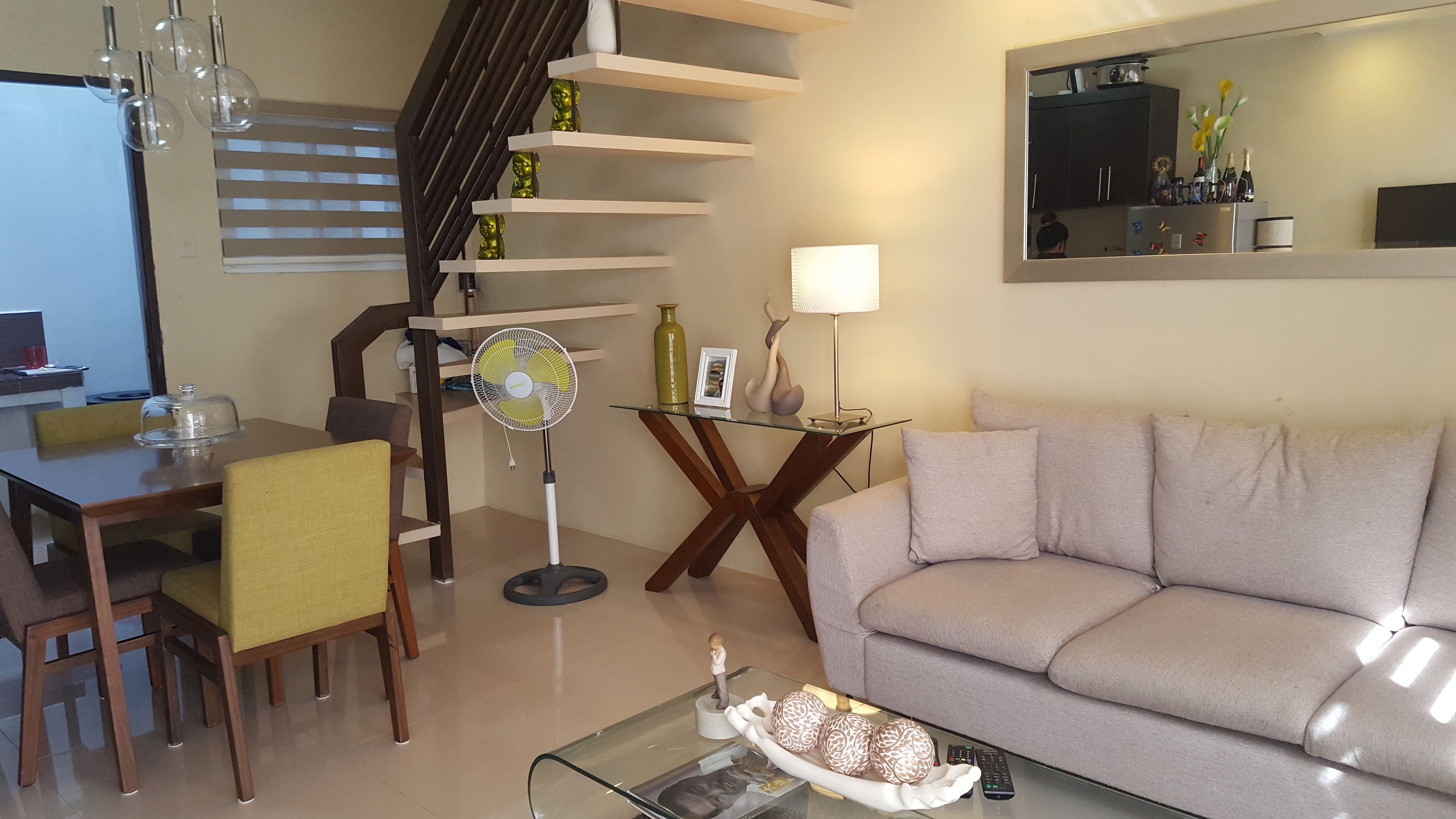 Un salon moderne comprenant un canapé, une table à manger, des chaises, une table basse en verre, un ventilateur sur pied et un escalier avec des étagères.
