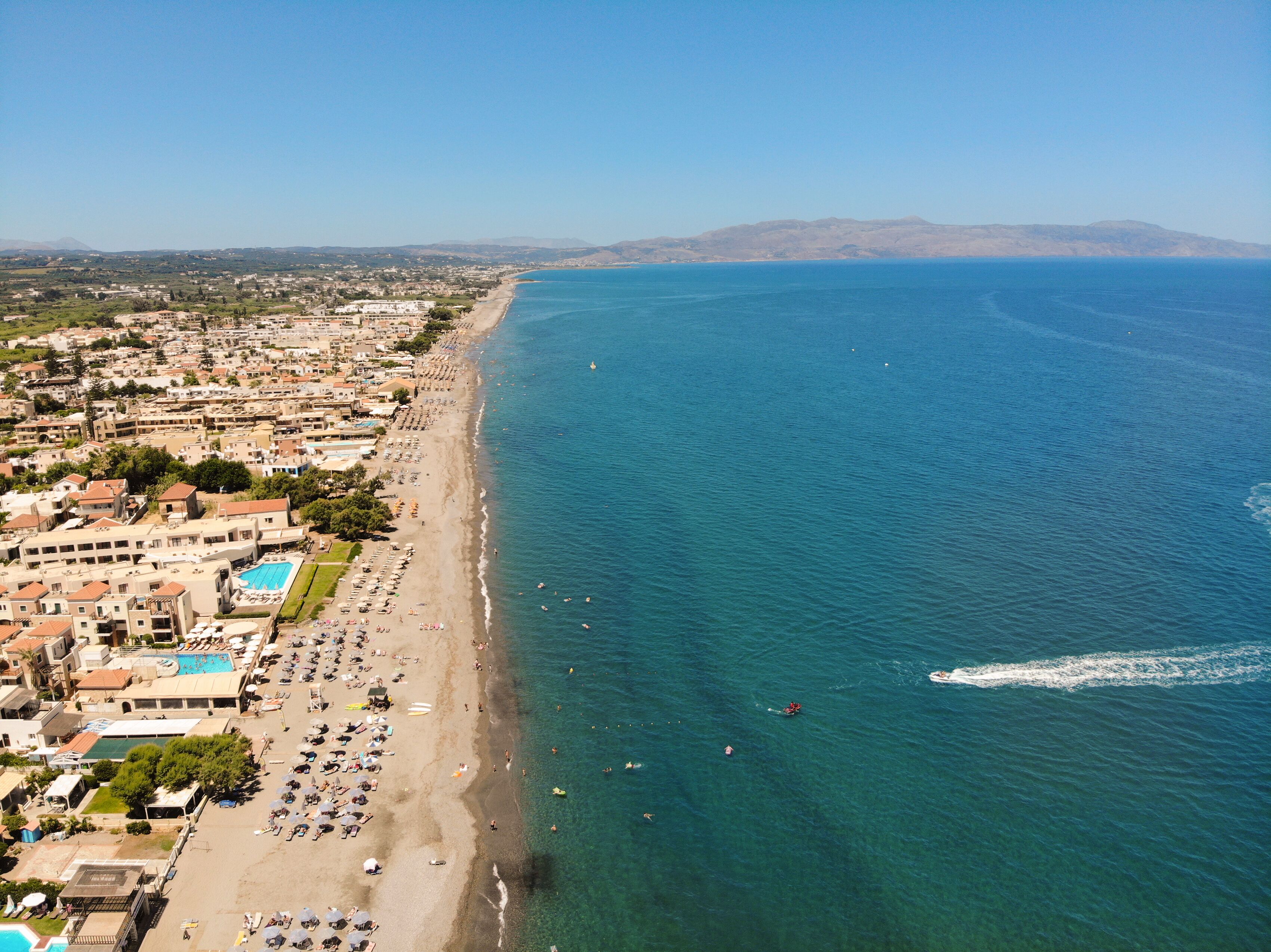 Vista aérea de un pueblo costero con una playa extensa, mar azul cristalino y montañas a lo lejos.