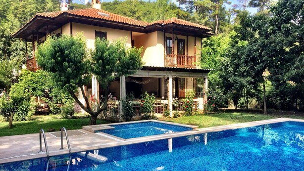 A poolside view of a house with a tiled pool, a covered patio, and lush greenery. - Defne Pansiyon (Marmaris)