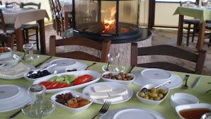 A dining table set with plates, bowls, and cutlery, surrounded by chairs, with a fireplace in the background. - Defne Pansiyon (Marmaris)