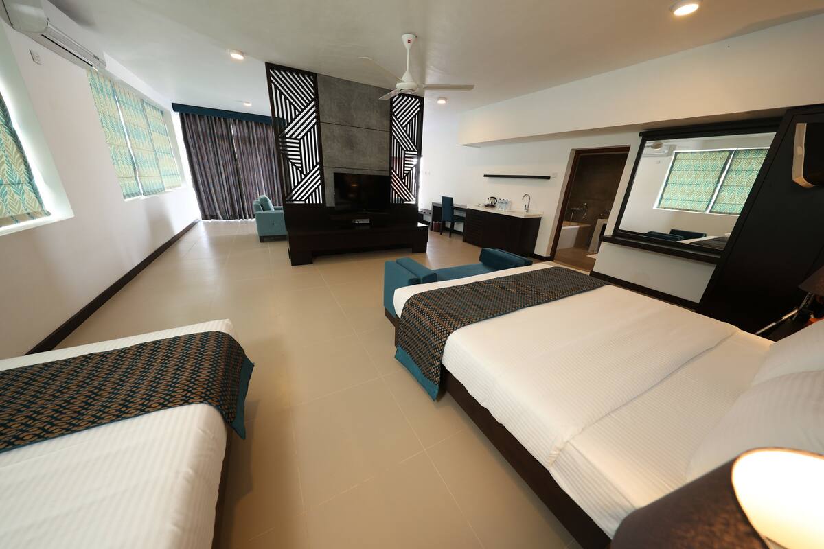 Habitación de hotel con dos camas, televisión, ventilador de techo, baño y zona de comedor.