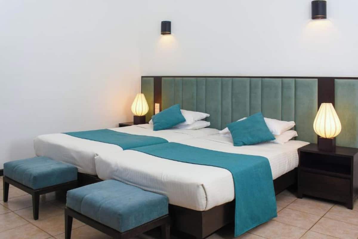 Una cama bien hecha con una colcha color turquesa y almohadas, flanqueada por dos mesitas de noche con lámparas, y un otoman a juego.