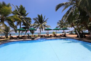 Un espace aménagé au bord de la piscine, avec des chaises longues et des parasols, des palmiers et une vue sur la plage.