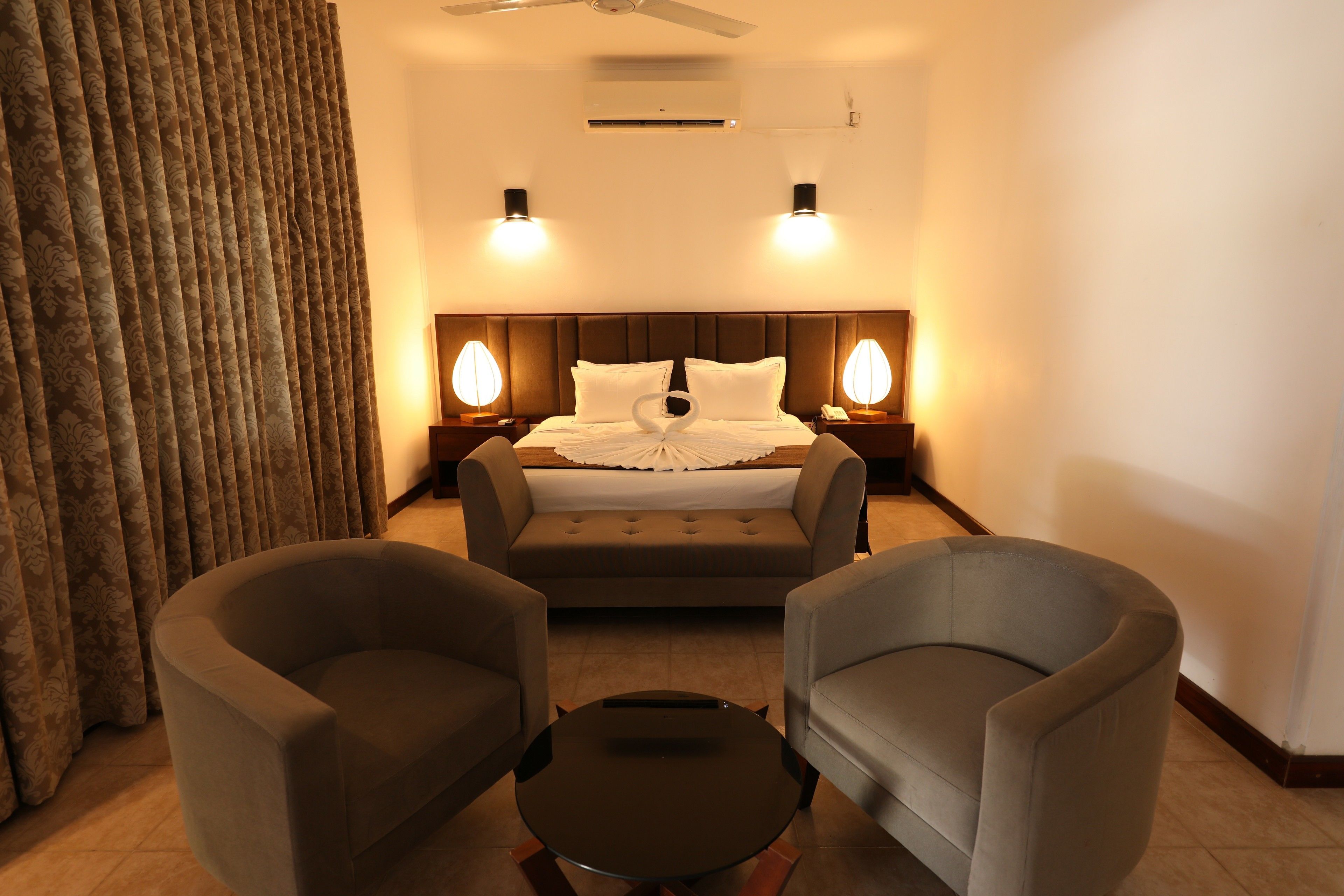 A hotel room with a bed, two bedside lamps, a ceiling fan, and two armchairs.