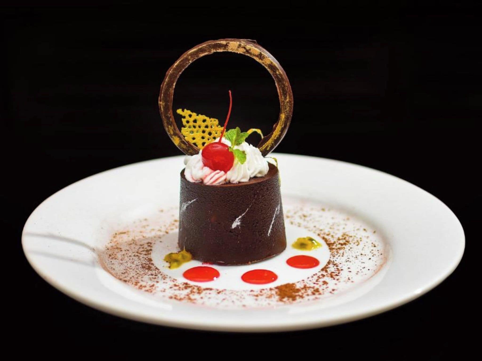 A dessert on a white plate with a chocolate cake, whipped cream, a cherry, and a decorative ring.