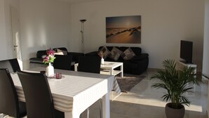 A modern living room with a dining area, a sofa set, a flat-screen TV, and a large painting on the wall. - 100 sqm large, central and quiet holiday home in Rhauderfehn! (Rhauderfehn)