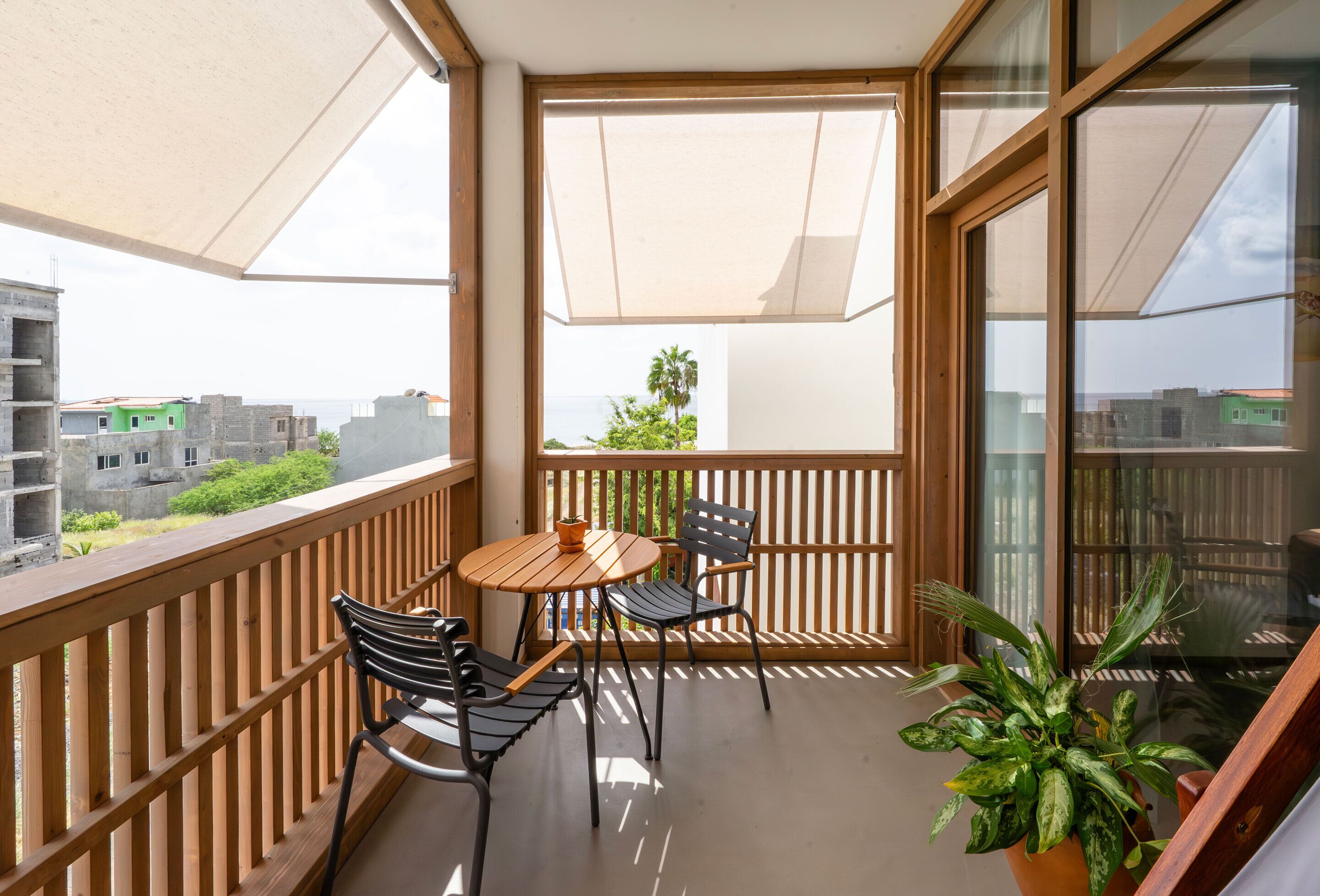 A balcony with a wooden table and chairs, a potted plant, and a view of buildings and greenery.