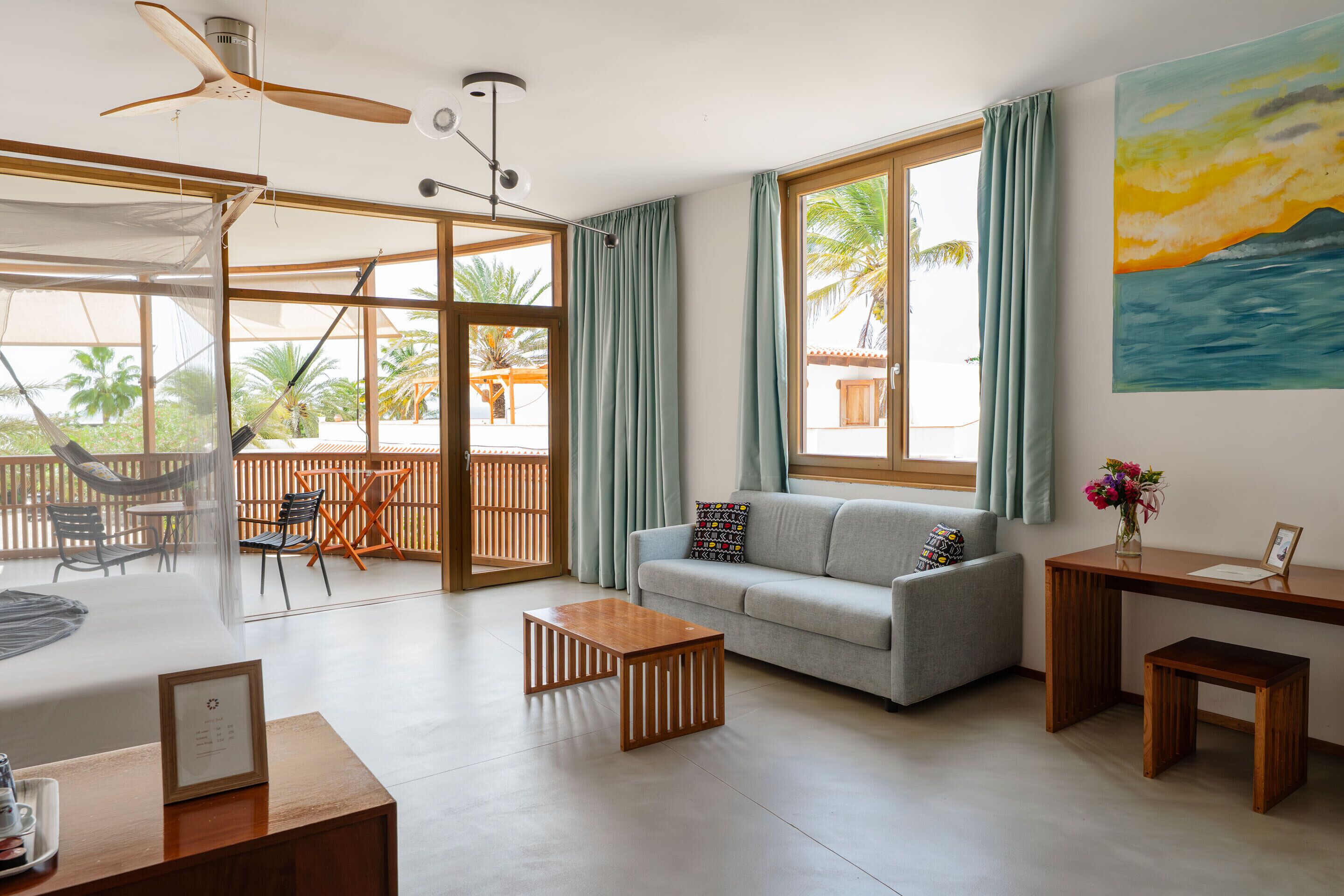 A spacious living area with a sofa, coffee table, and a desk. There is a large window with curtains, a ceiling fan, and a painting on the wall.