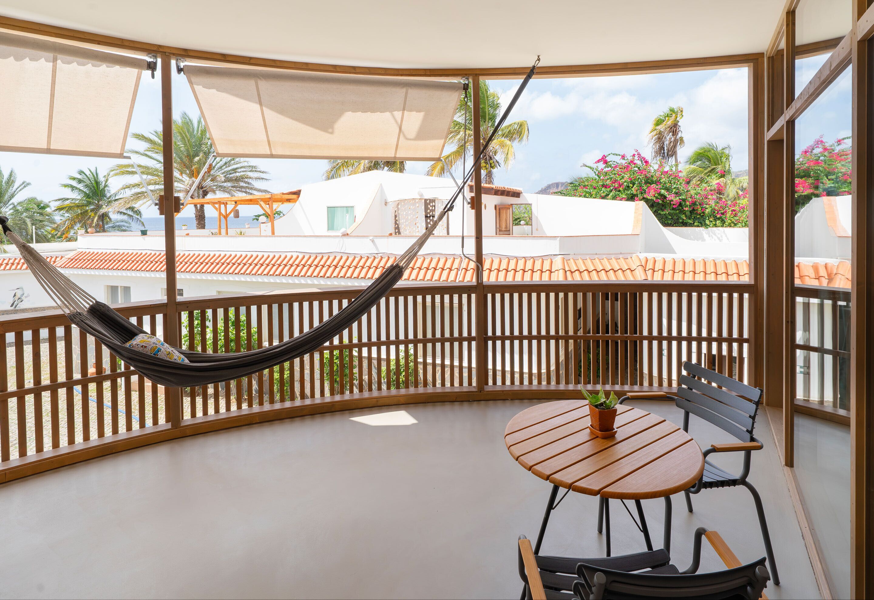 A balcony with a hammock, a wooden table with chairs, and a view of buildings with red-tiled roofs.
