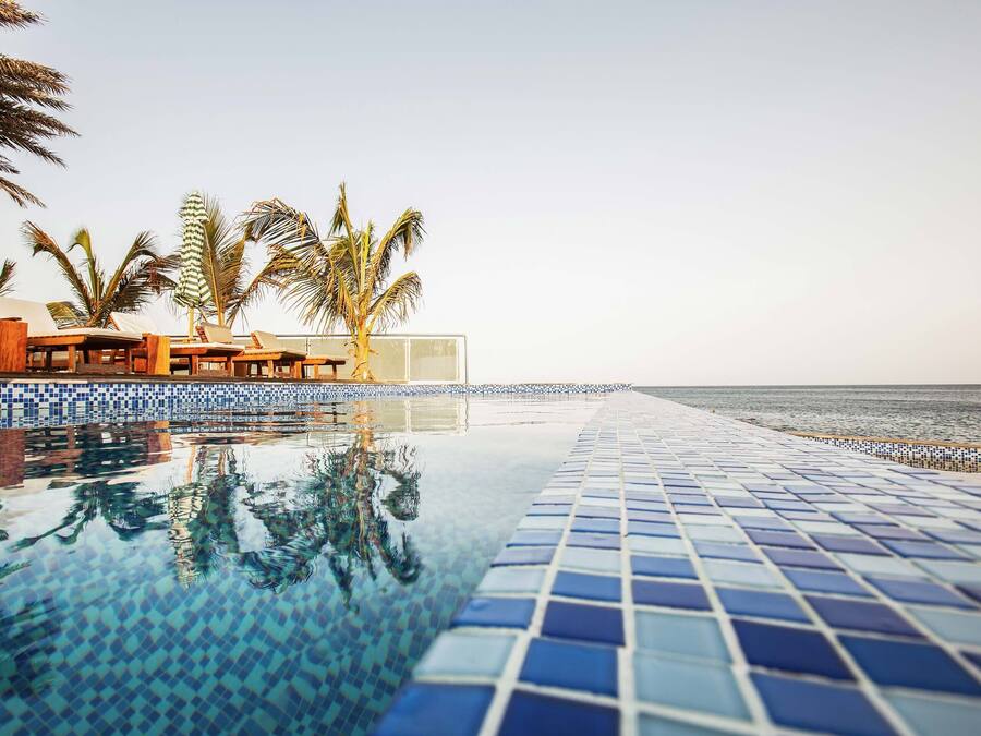 A pool with blue mosaic tiles, lounge chairs, and palm trees.
