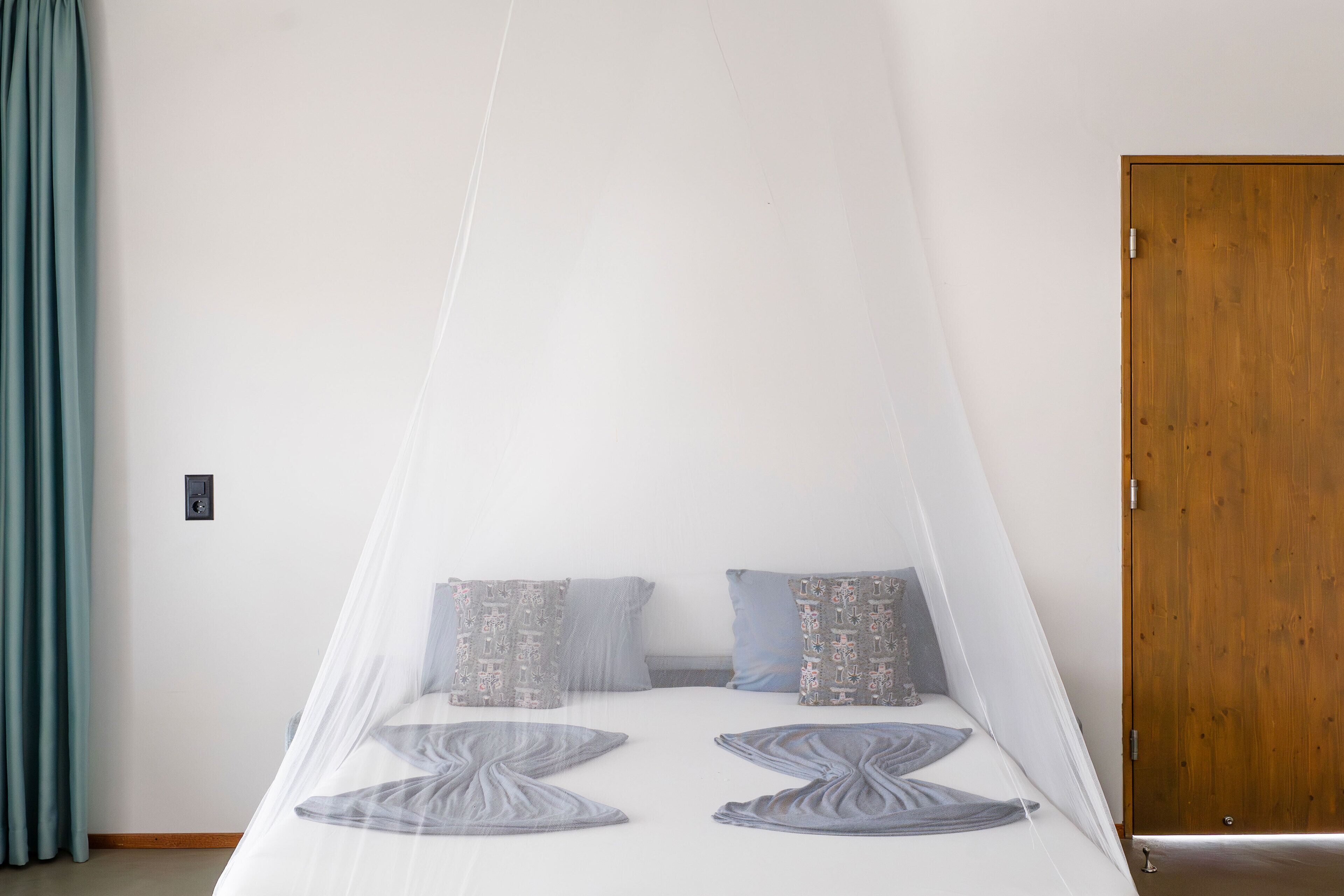 Une chambre à coucher avec un lit recouvert d’une moustiquaire, deux coussins à motifs, une porte en bois et un rideau de couleur turquoise.