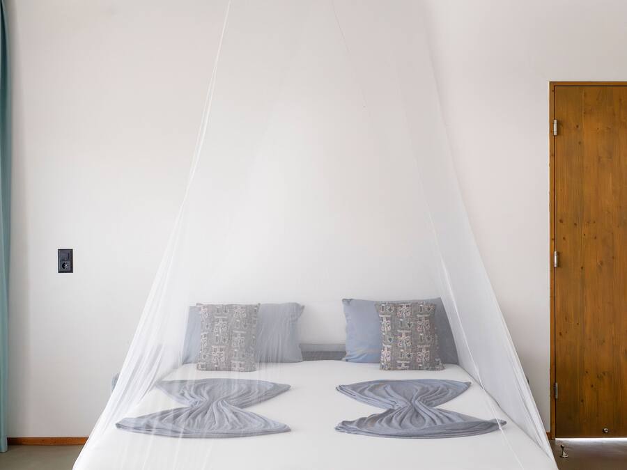 Une chambre à coucher avec un lit recouvert d’une moustiquaire, deux coussins à motifs, une porte en bois et un rideau de couleur turquoise.