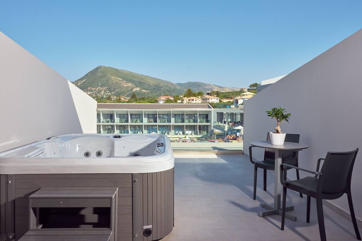 A rooftop terrace with a hot tub, a small table with chairs, and a potted plant. In the background, there is a building with multiple floors and a mountain view.