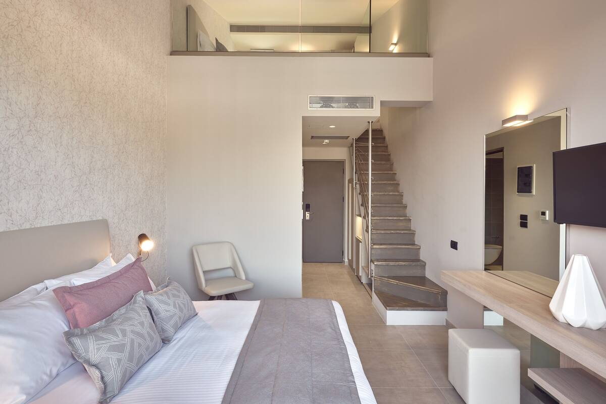 A modern hotel room with a large bed, a chair, a desk, a mirror, and a staircase leading to an upper level.