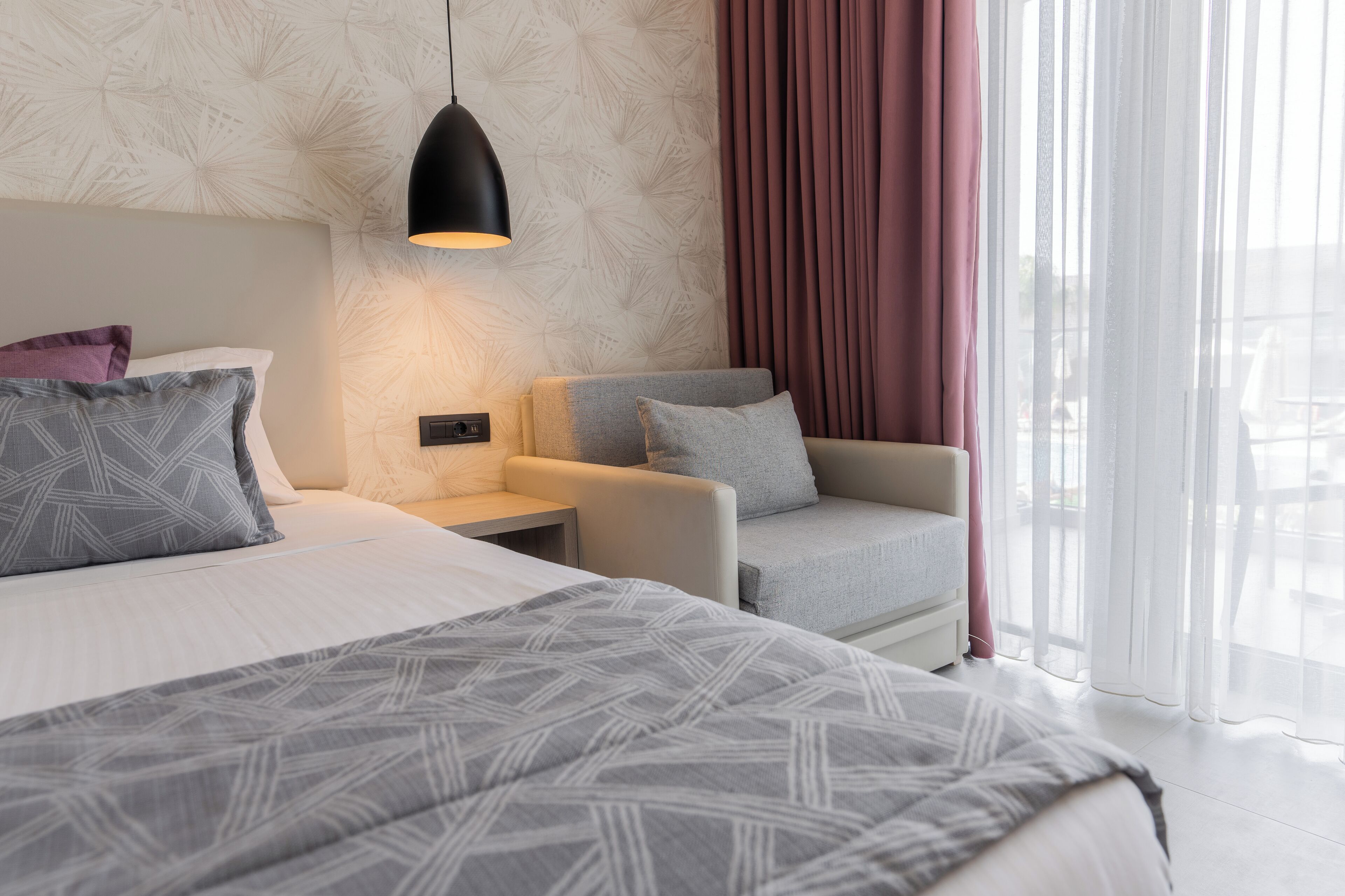 A modern hotel room with a large bed, a seating area with a chair, a nightstand, and a wall-mounted lamp.