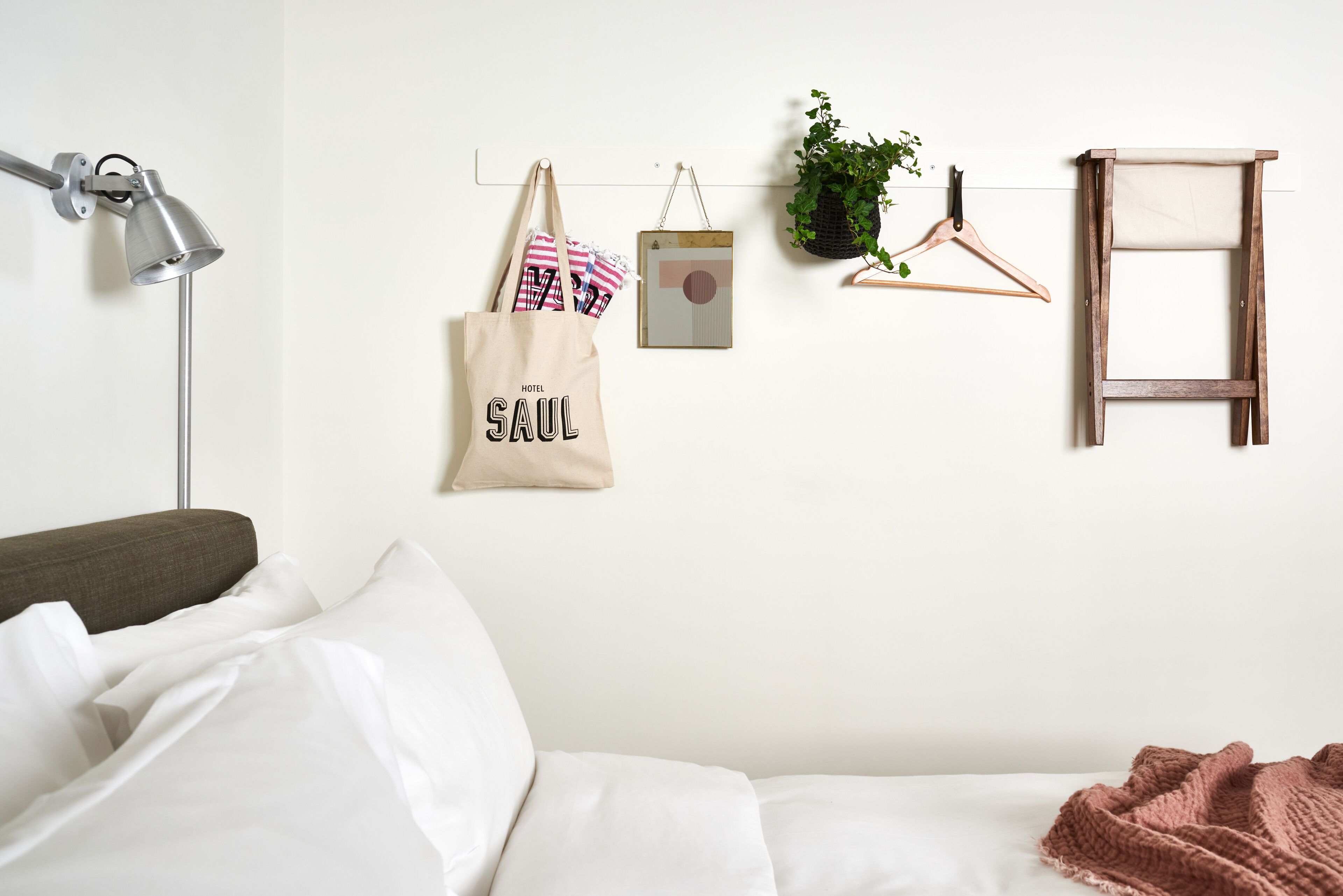 A neatly made bed with white bedding, a grey floor lamp, a hanging plant, a wooden hanger, and a beige tote bag with "HOTEL SAUL" text.