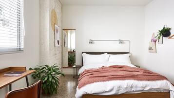 Quarto com cama de madeira, roupa de cama branca, escrivaninha com cadeira, planta em vaso e janela com persianas.