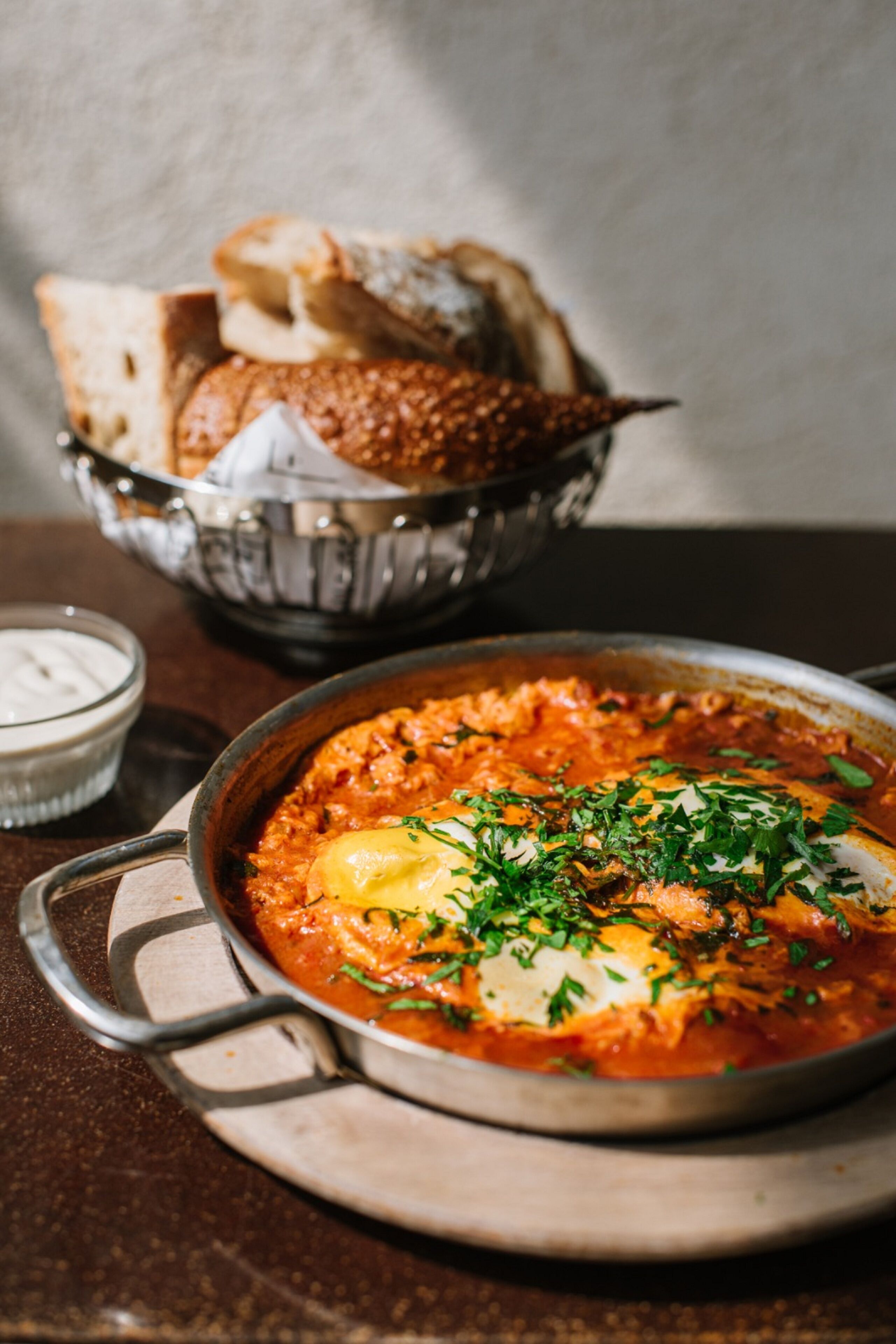 A bowl of shakshuka with poached eggs and herbs, accompanied by a basket of bread and a small bowl of yogurt.