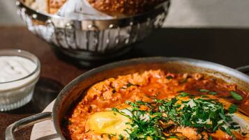 Un tazón de shakshuka con huevos escalfados y hierbas, acompañado de una cesta de pan y un pequeño tazón de yogur.