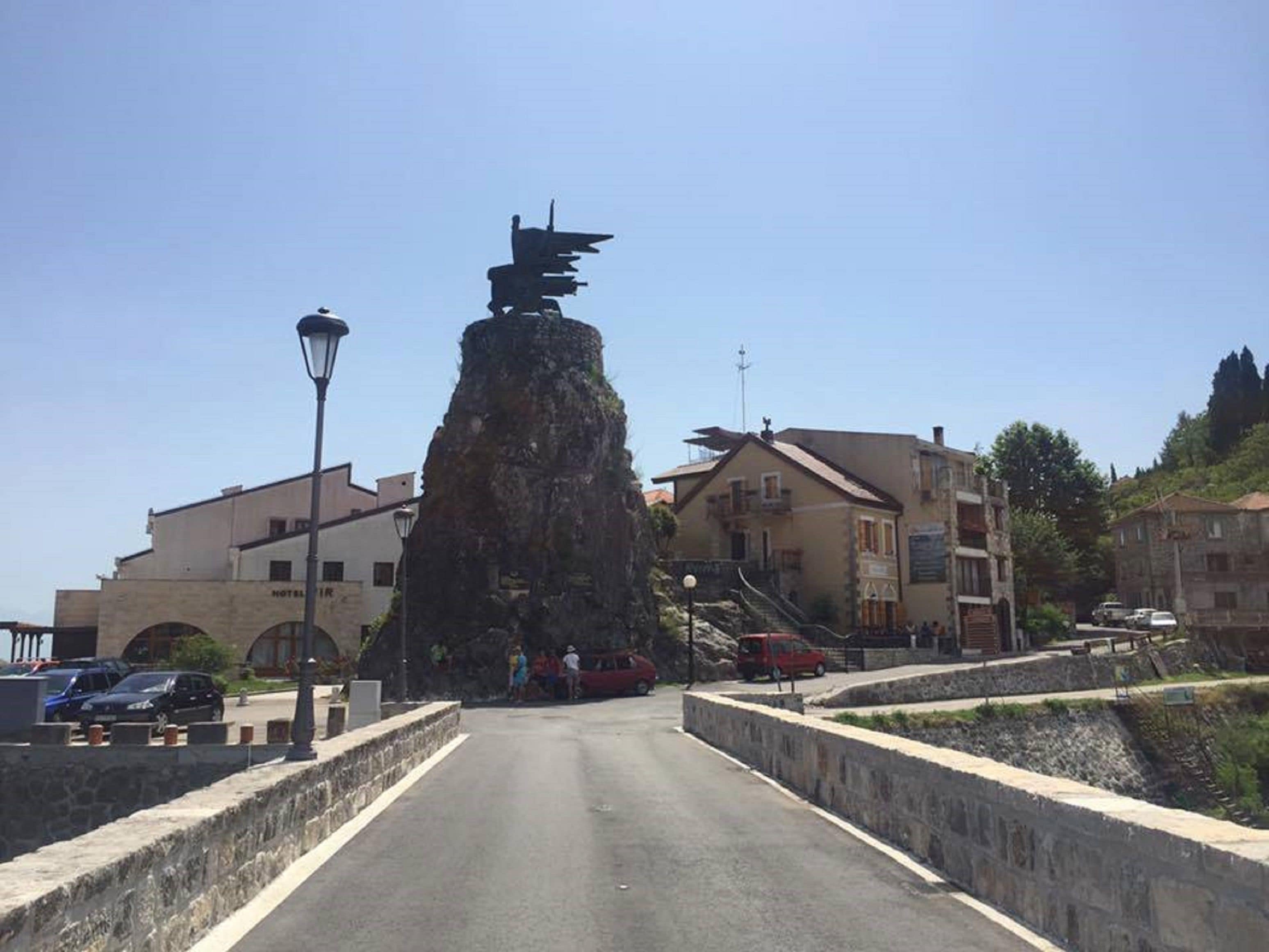 A street with a prominent rock formation in the center, surrounded by buildings and a clear sky.
