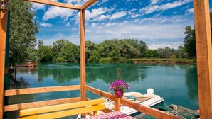 Een houten terras met een bank en tafel, met uitzicht op een kalme rivier met boten en begroeiing op de achtergrond.