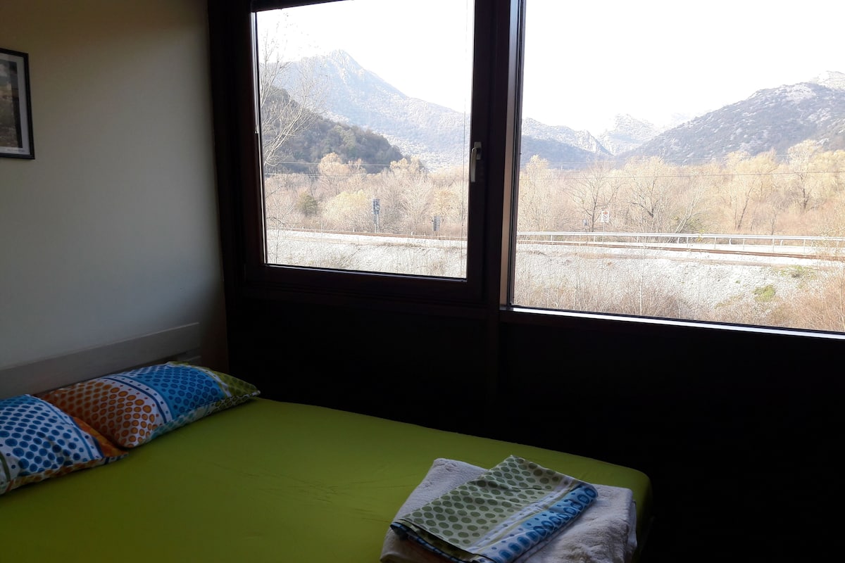 Una stanza con un letto, cuscini e una finestra vista montagne e binari ferroviari.