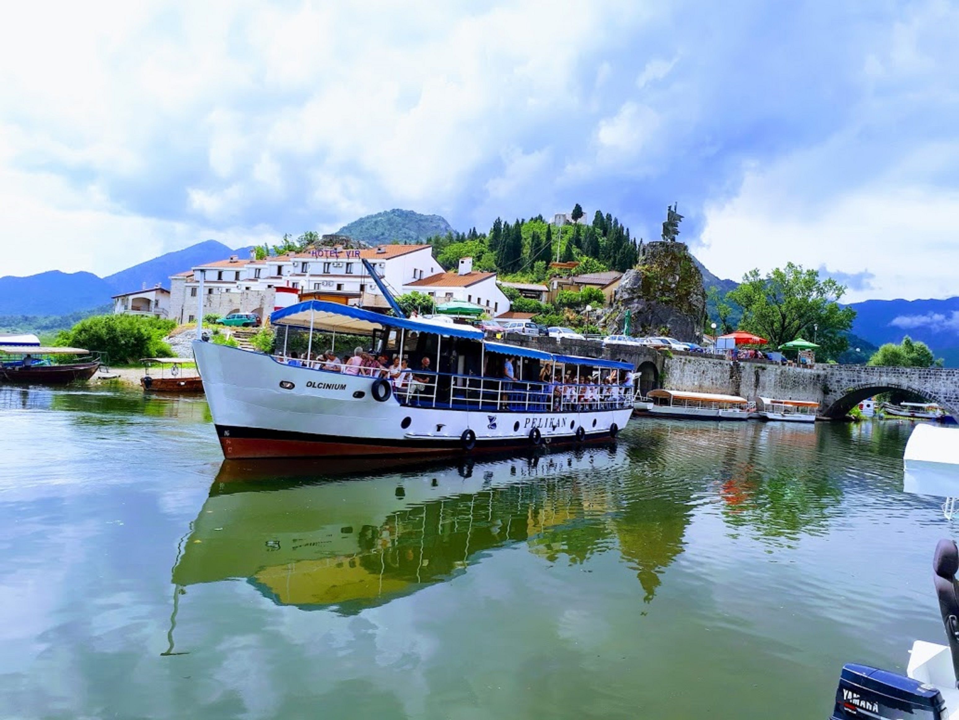 A boat with the name "PELIKAN" docked at a riverside location with a bridge and buildings in the background.