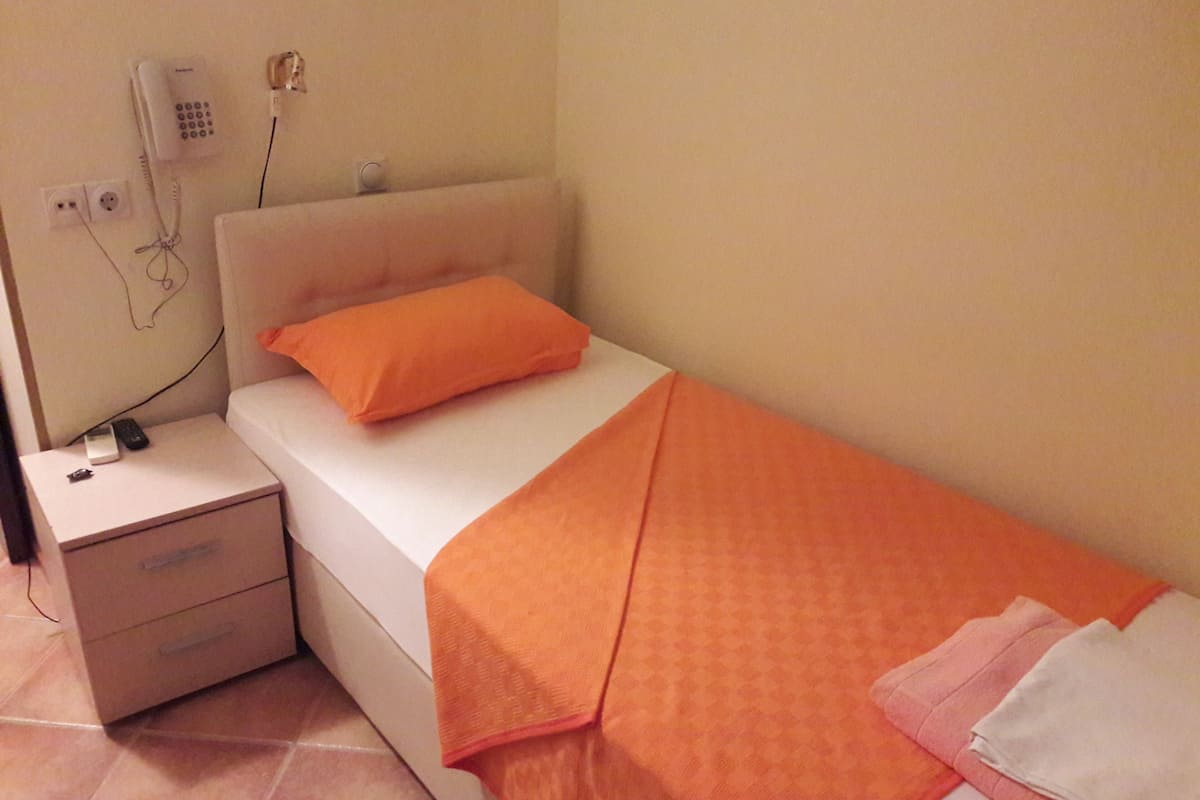 Un letto singolo con coperta e cuscino arancioni, un comodino con telefono e telecomando, e una presa elettrica a muro.