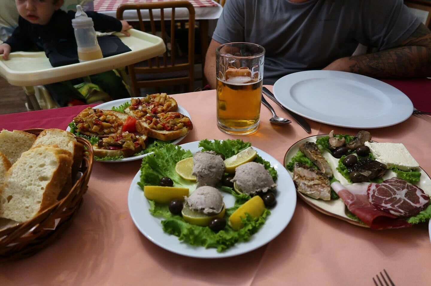 Una mesa con diversos platos, una cerveza en un vaso y un bebé en una trona.