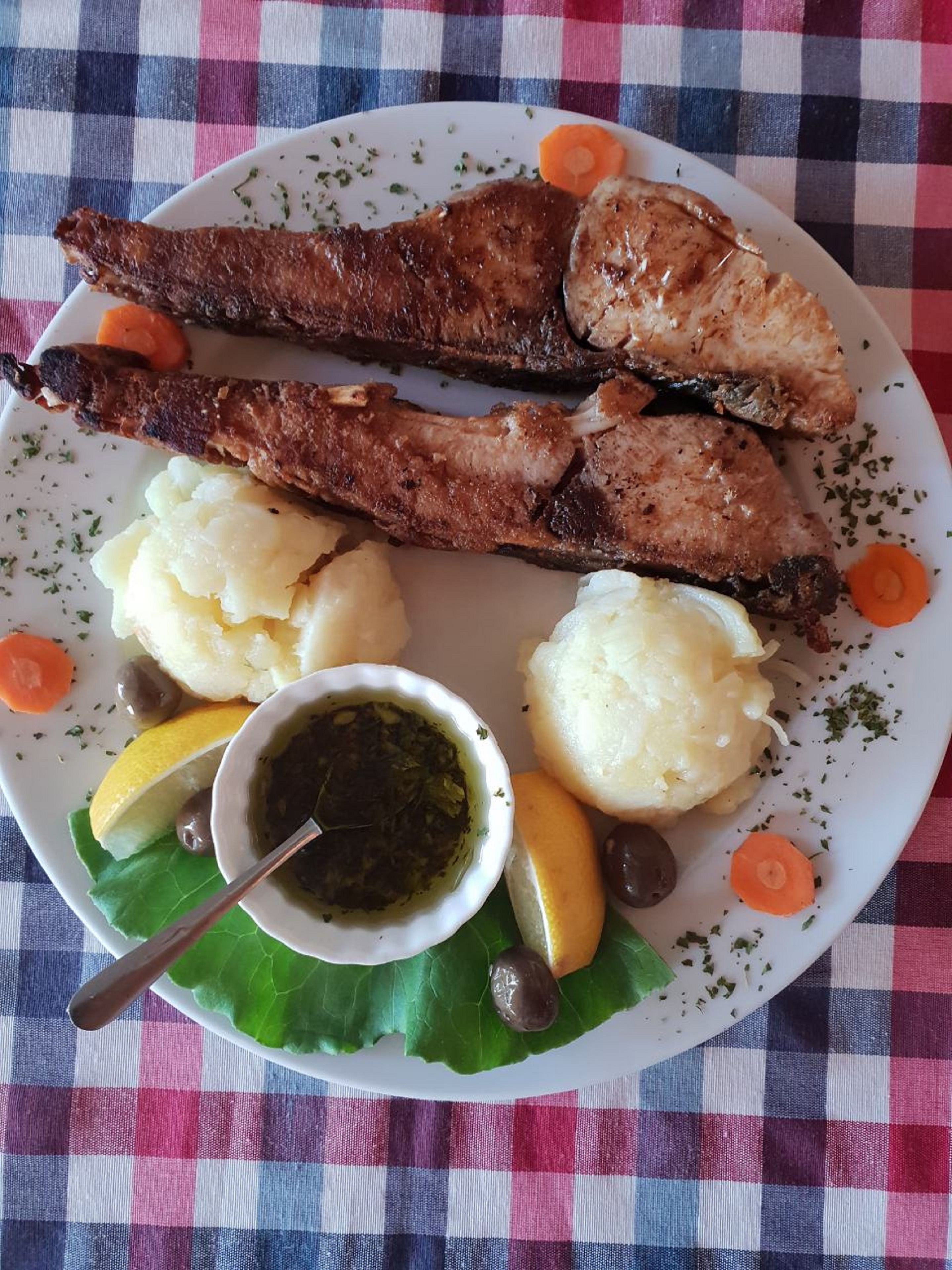 Un plato de comida que incluye carne asada, puré de papas, una rodaja de limón, aceitunas y un pequeño recipiente con salsa, sobre un mantel a cuadros.