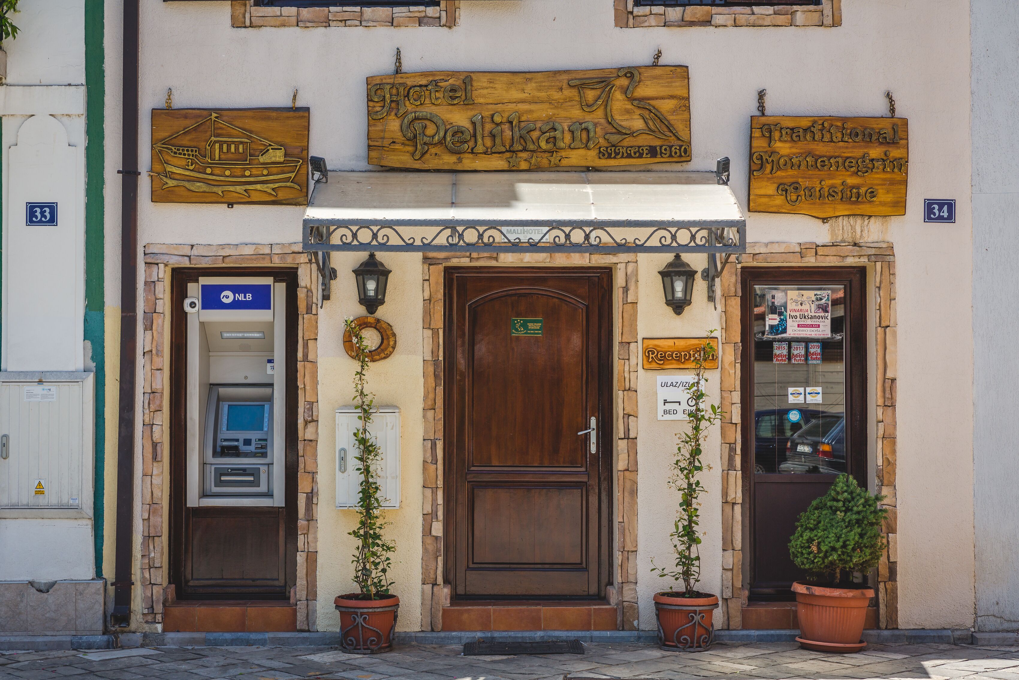 A hotel entrance with a wooden signboard, an ATM, and two potted plants.