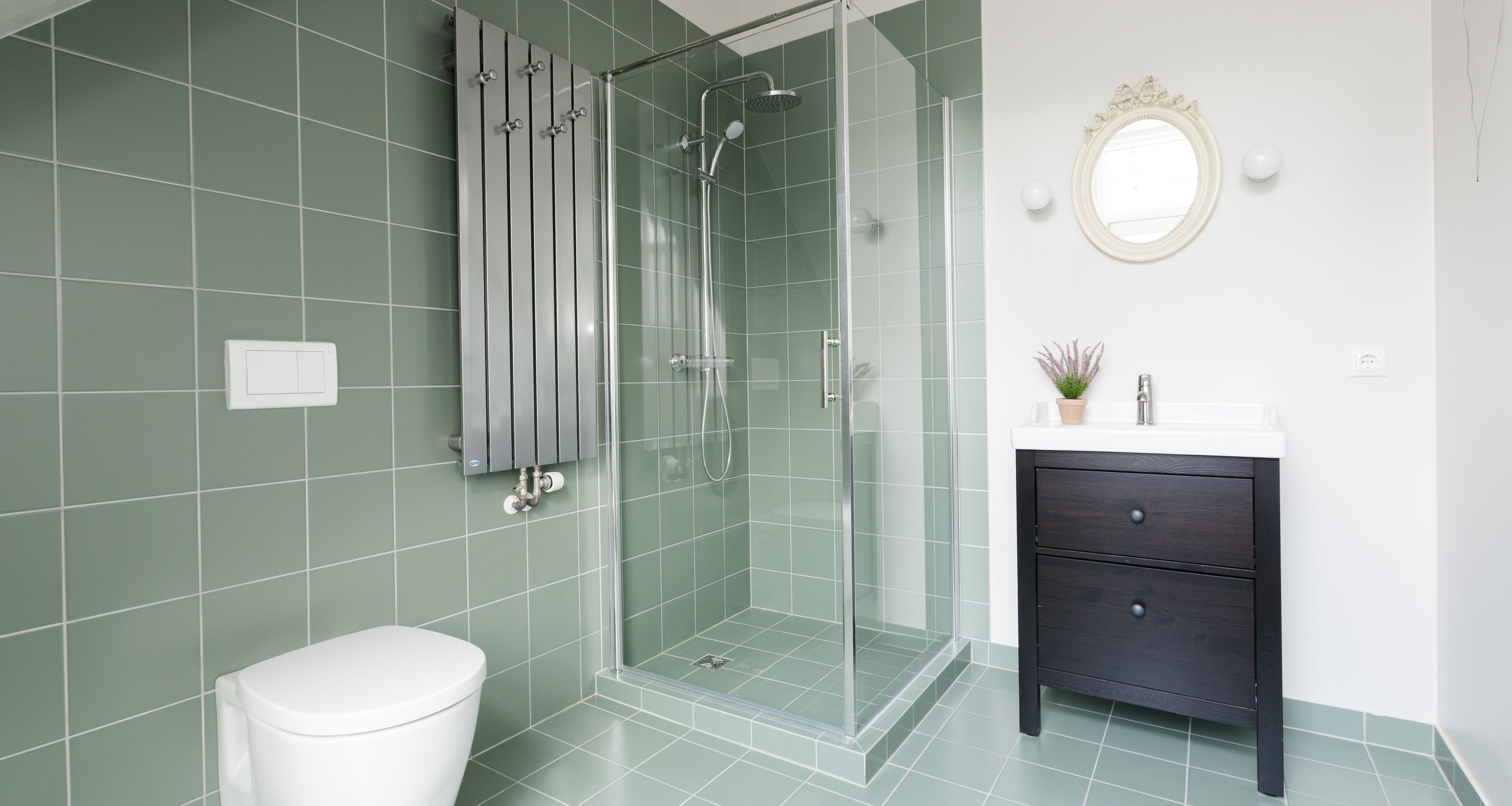 A bathroom with a glass-enclosed shower, a white toilet, a dark wooden vanity with a mirror, and green tiled walls.