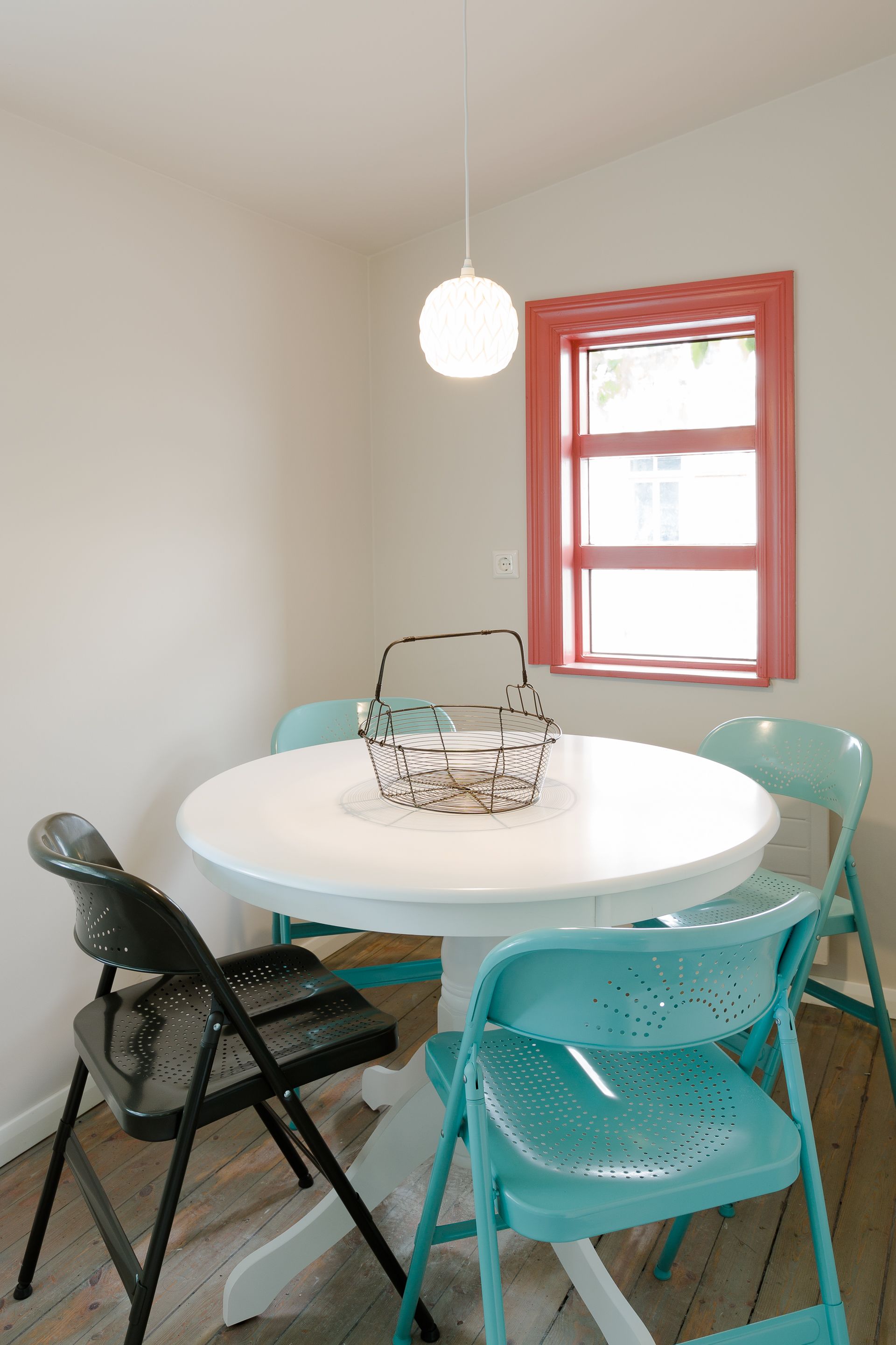 A dining area with a round white table, teal chairs, a black chair, a wire basket, and a hanging light fixture.