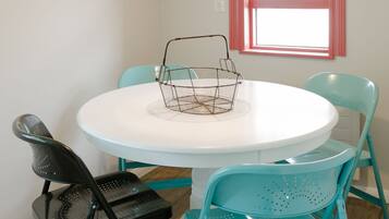 A dining area with a round white table, teal chairs, a black chair, a wire basket, and a hanging light fixture.