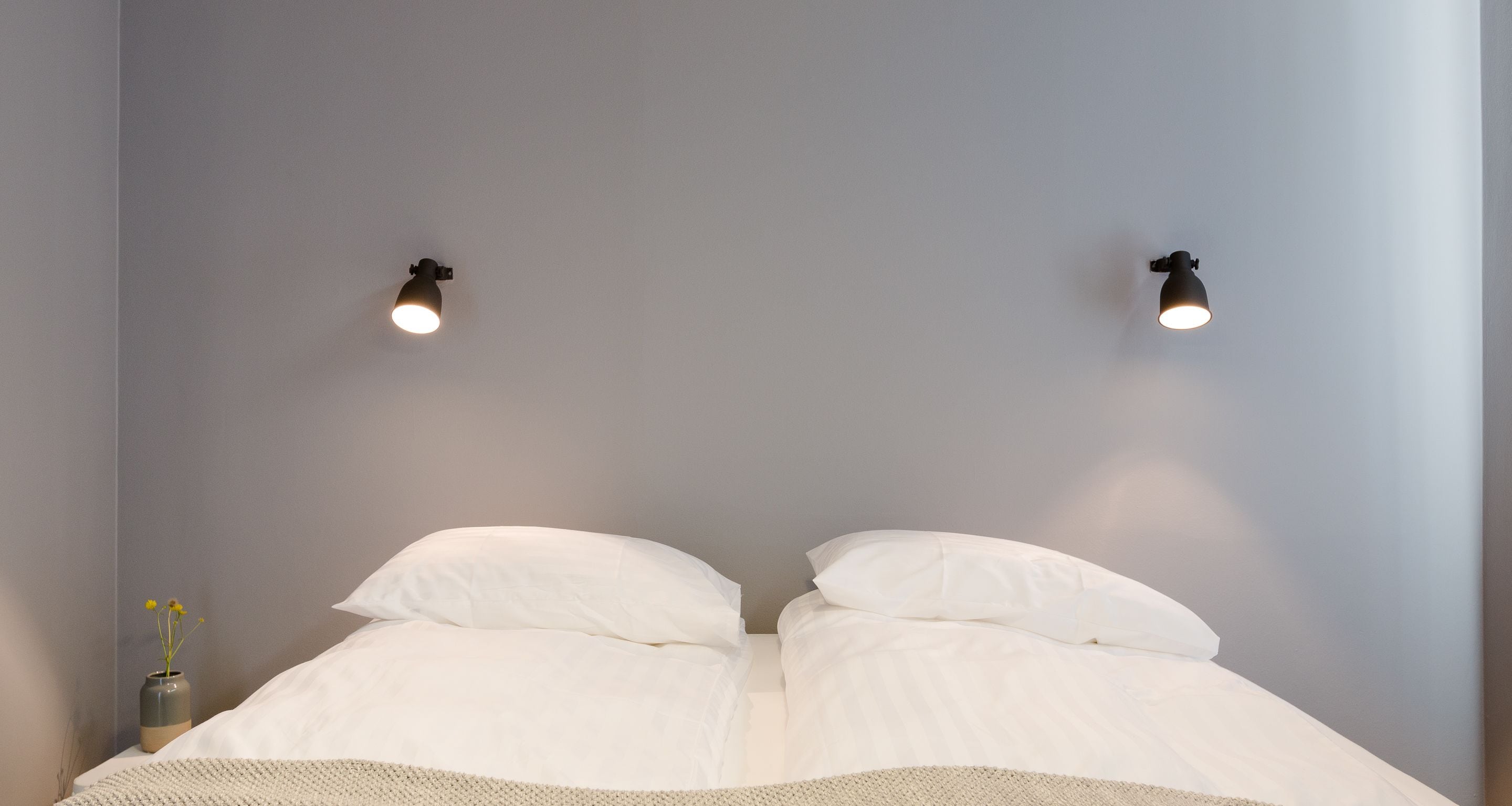 Une chambre à coucher avec un lit, deux lampes fixées au mur et un petit vase avec des fleurs sur la table de chevet.