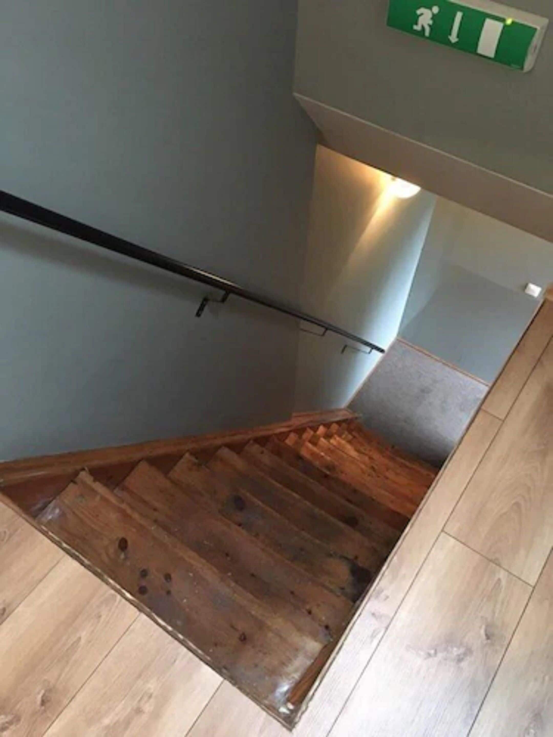Un escalier en bois avec une rampe, un panneau de signalisation verte indiquant la sortie et un luminaire au-dessus.