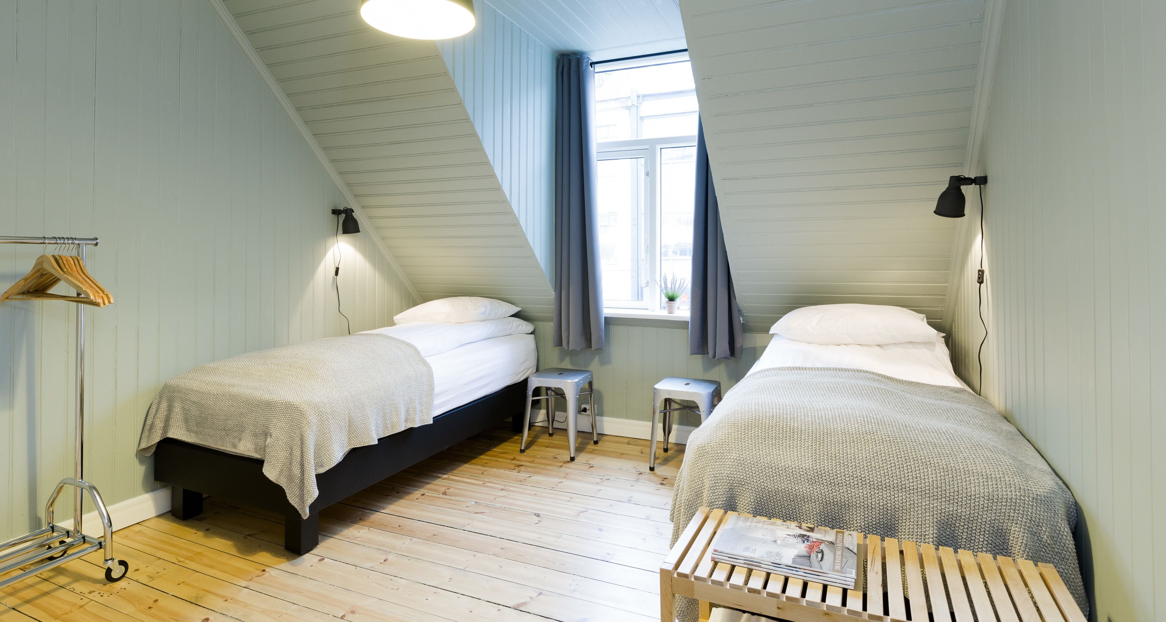 Une chambre avec deux lits, une petite table avec un magazine, un porte-vêtements et une fenêtre avec des rideaux.