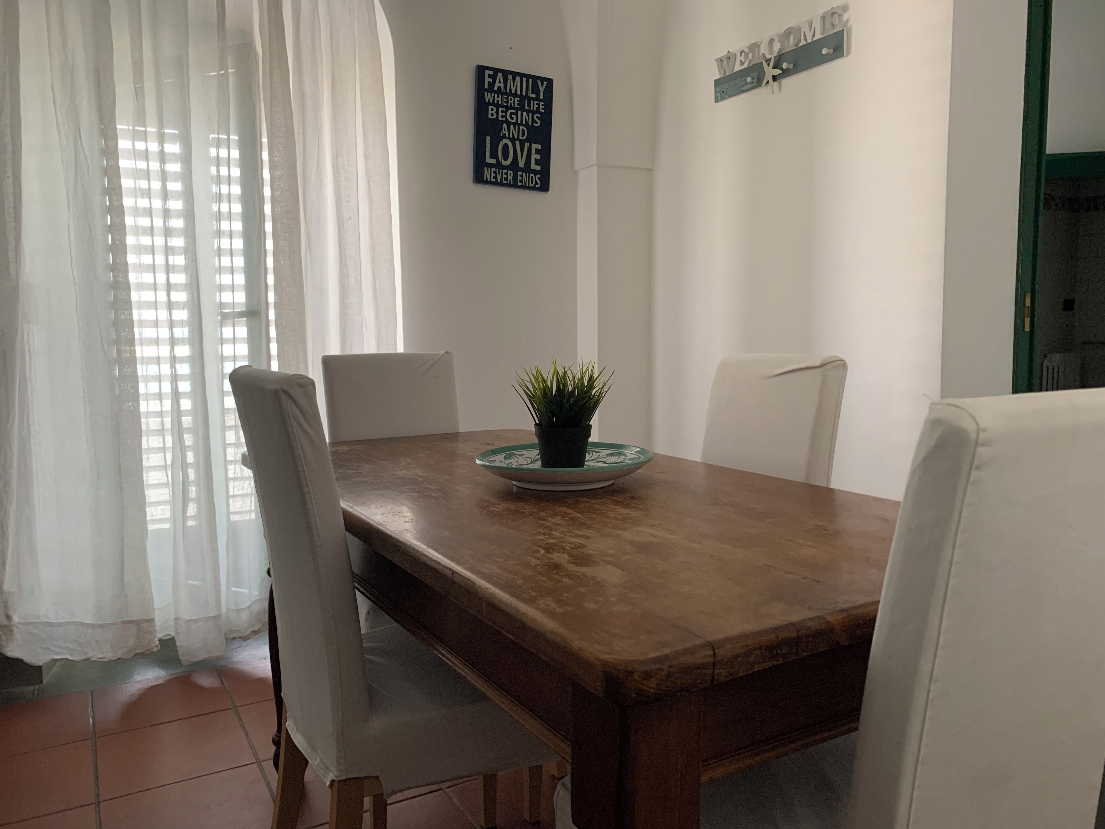 Ein Essbereich mit einem Holztisch, weißen Stühlen, einer Kü Pflanze und einem 'WELCOME'-Schild an der Wand.