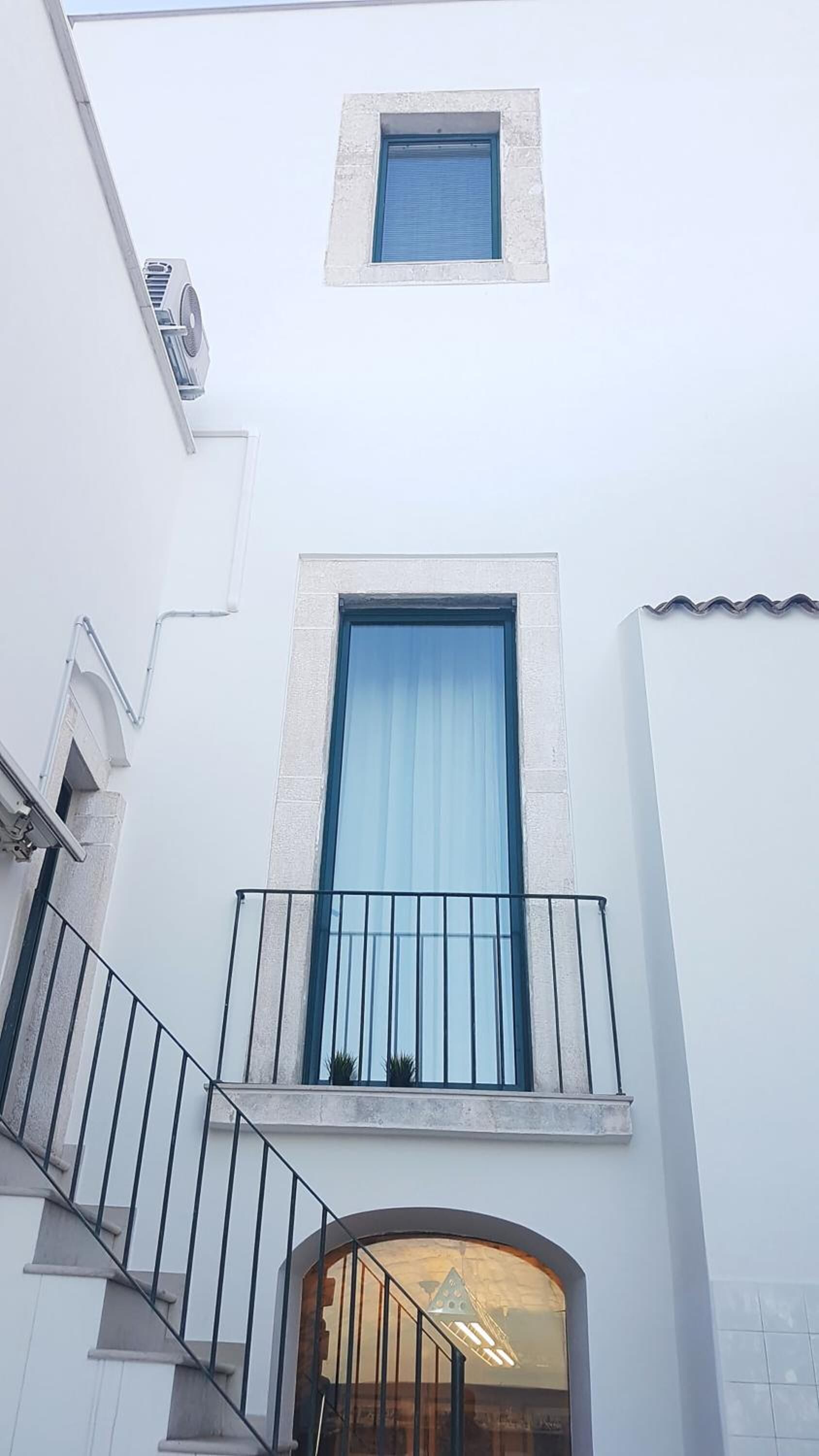 Un edificio con un ventanal grande, un ventanuilla más pequeña arriba y una escalera que conduce a un balcón con barandillas.