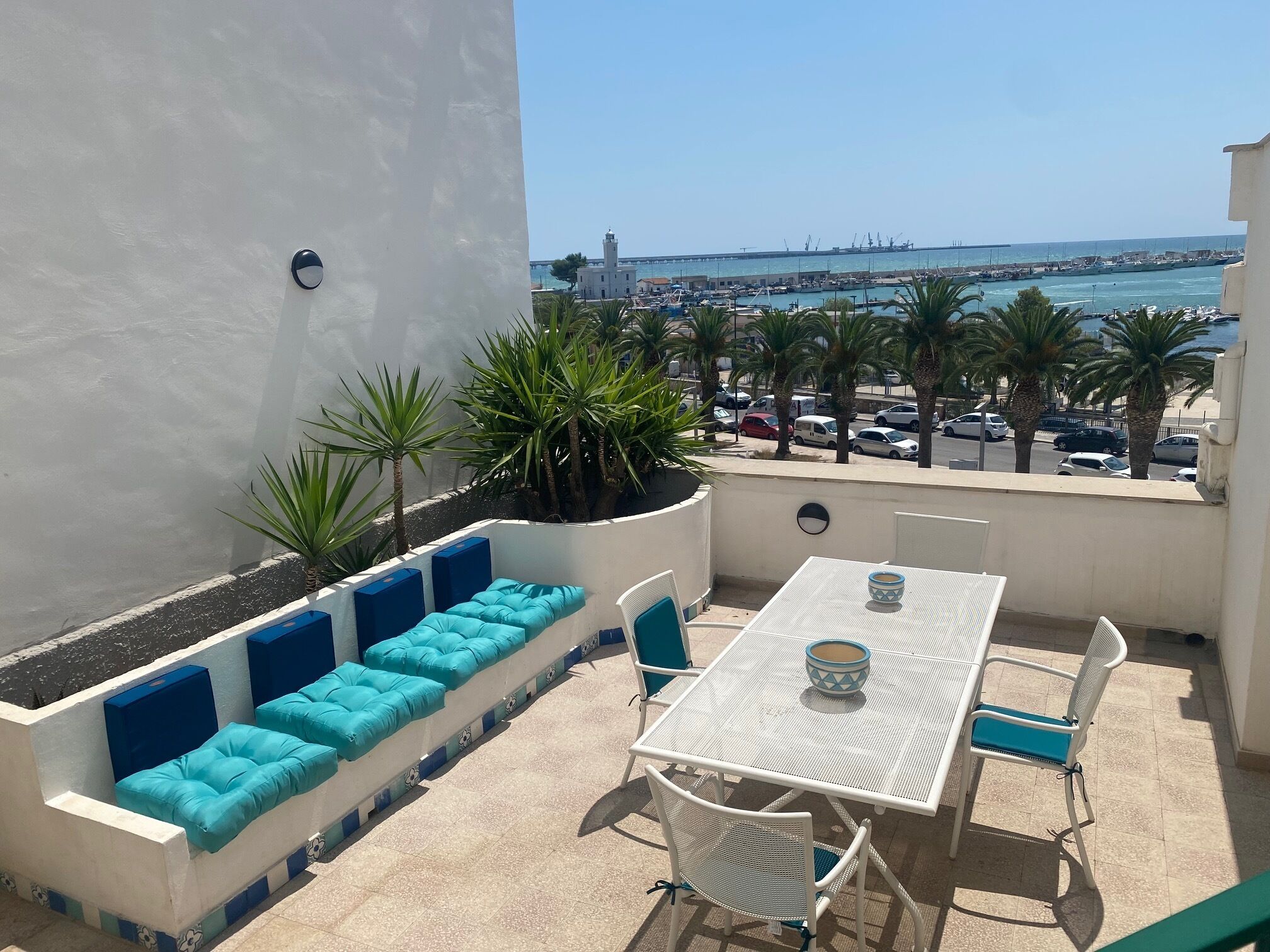 A balcony with a white table and chairs, a blue cushioned bench, and a view of the sea and port.
