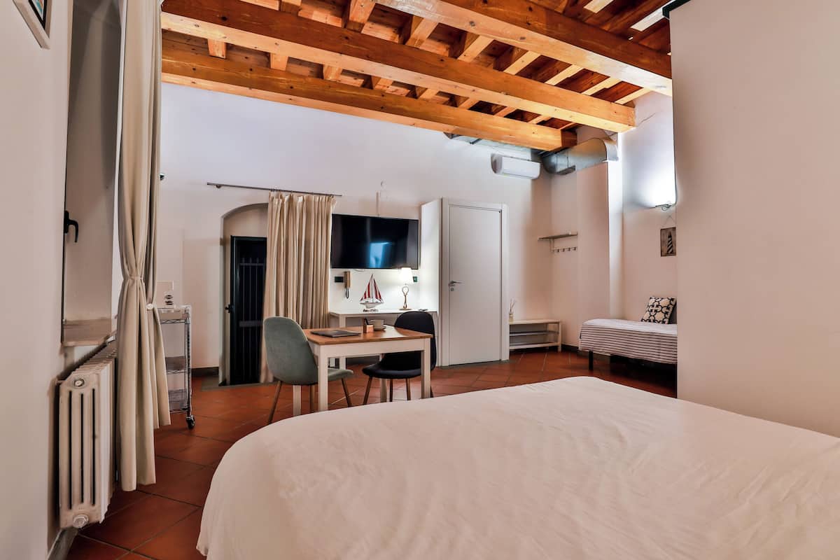 A bedroom with a bed, a desk, a chair, a TV, and a door leading to another room.