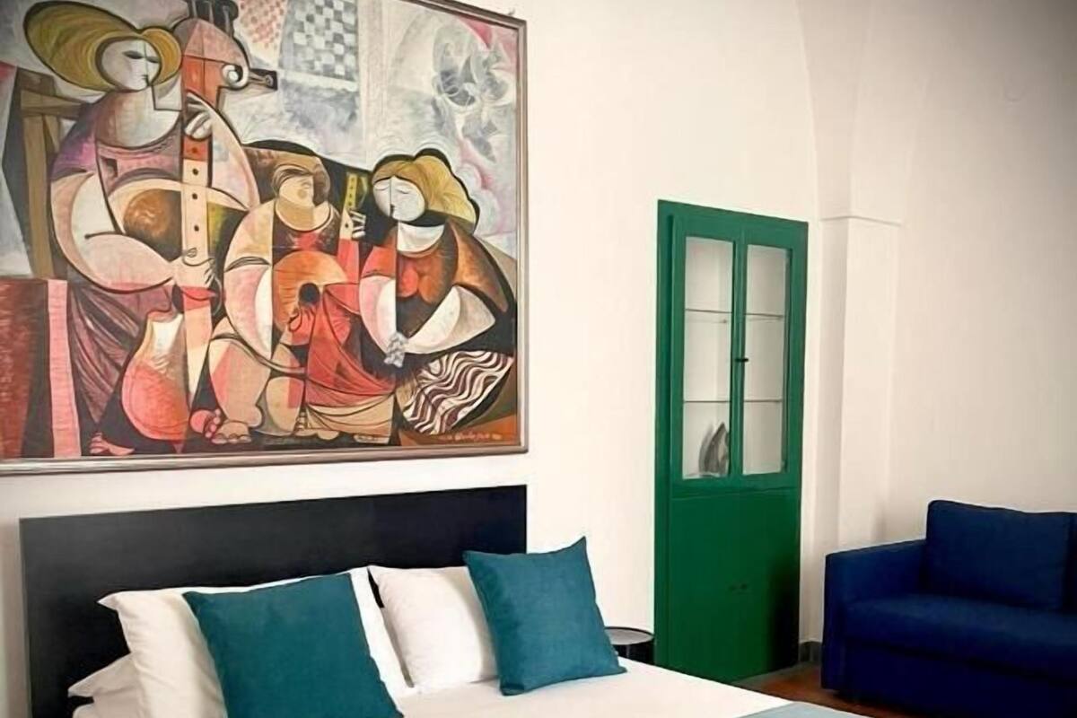 A bedroom with a bed, a blue chair, and a large abstract painting on the wall.
