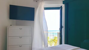 Un dormitorio con cama, cómoda blanca, televisor montado en la pared y balcón con vista al mar.